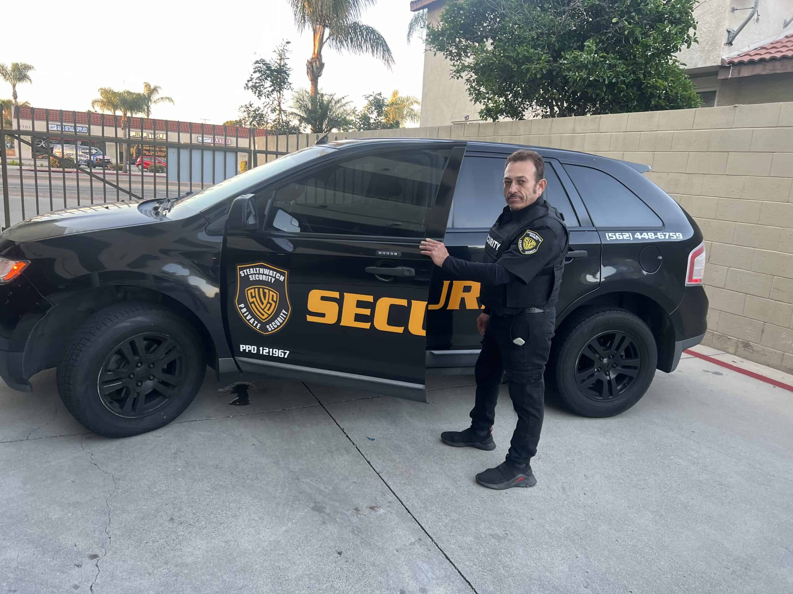 An armed security guard stands next to a black security vehicle with the word "SECURITY" written on the side in yellow letters. The guard is holding the driver-side door handle with his left hand. The scene is set in an outdoor, urban area in Los Angeles County, with palm trees visible in the background.