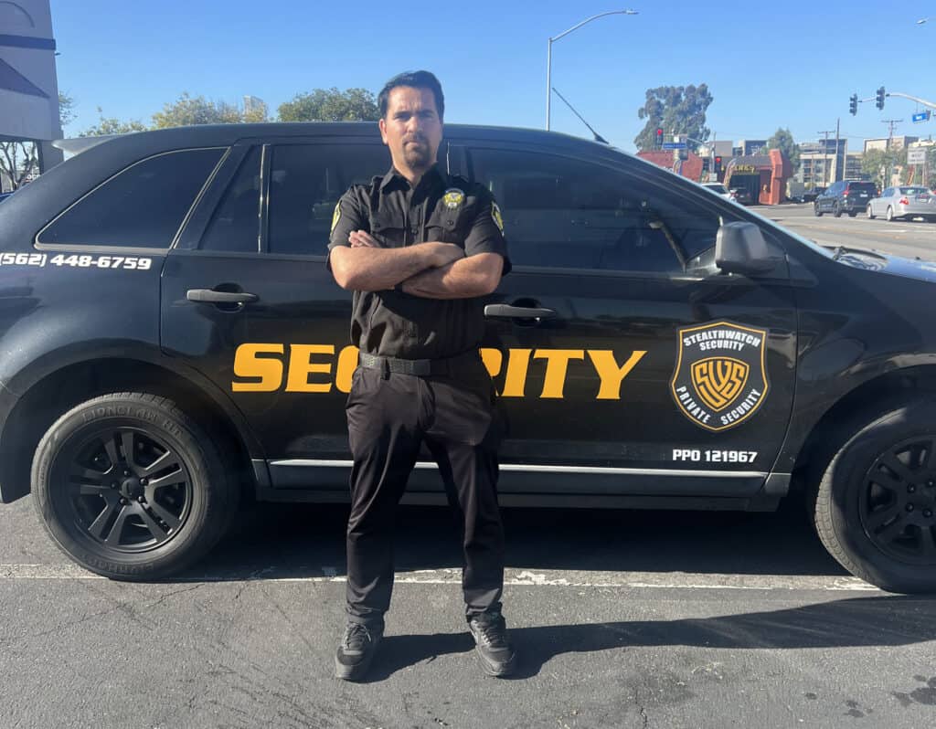 A security officer stands with arms crossed in front of a black SUV labeled "SECURITY" on the side. The officer, an Armed Security Guard Los Angeles County professional, wears a black uniform. In the background, there is a street with some cars and buildings visible under a clear sky.