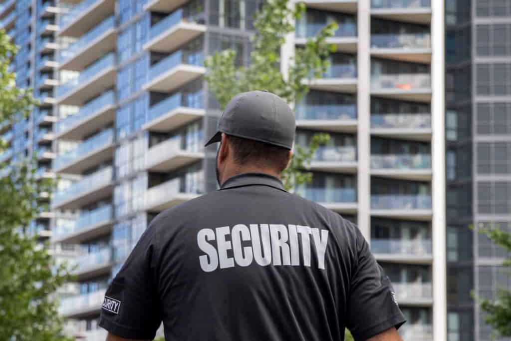 A security guard in uniform with "SECURITY" written on the back is standing in front of a modern high-rise building. The building features glass balconies, and there are green trees in the foreground.
